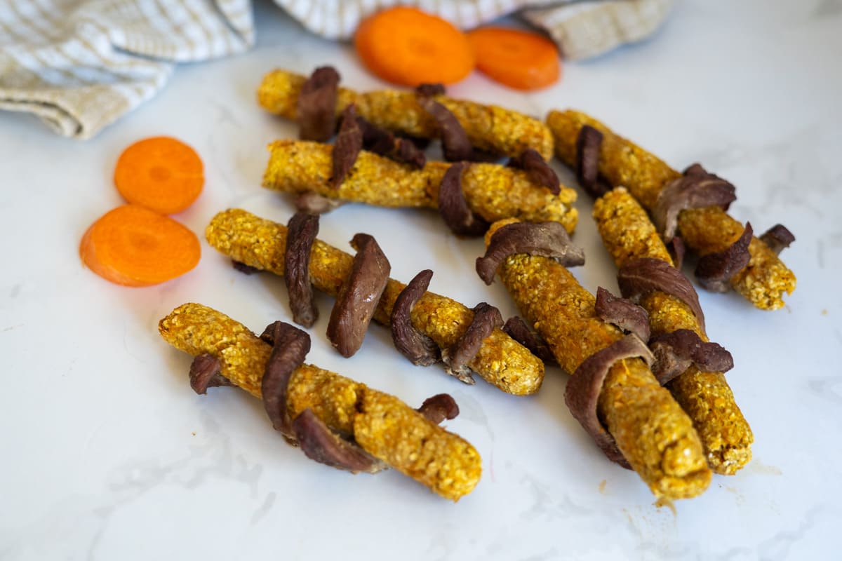 Several homemade dog treats wrapped in dried meat are arranged on a white surface, with carrot slices and a cloth in the background.