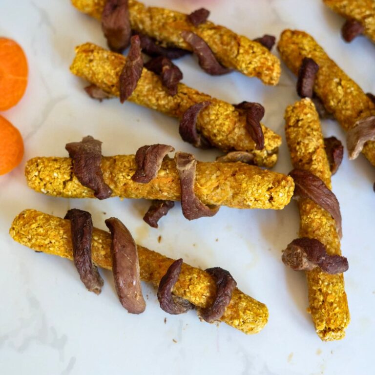 Dog treats made from oat and carrot sticks wrapped with pieces of dried meat arranged on a white surface.