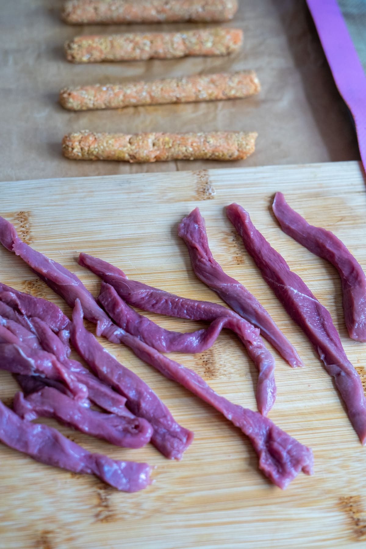 Raw meat slices on a wooden chopping board with several homemade duck dog treats and breaded sticks on baking paper in the background.