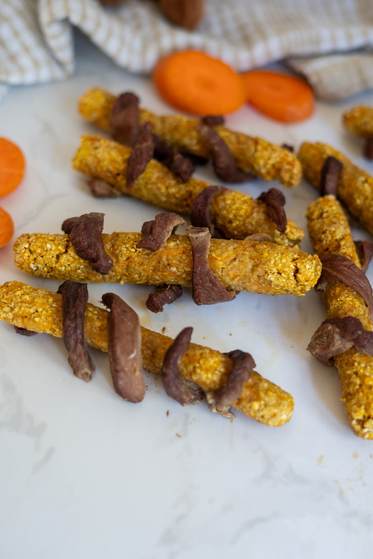 Homemade duck dog treats shaped like sticks, wrapped with strips of dried meat, are arranged on a white surface with slices of carrot in the background.