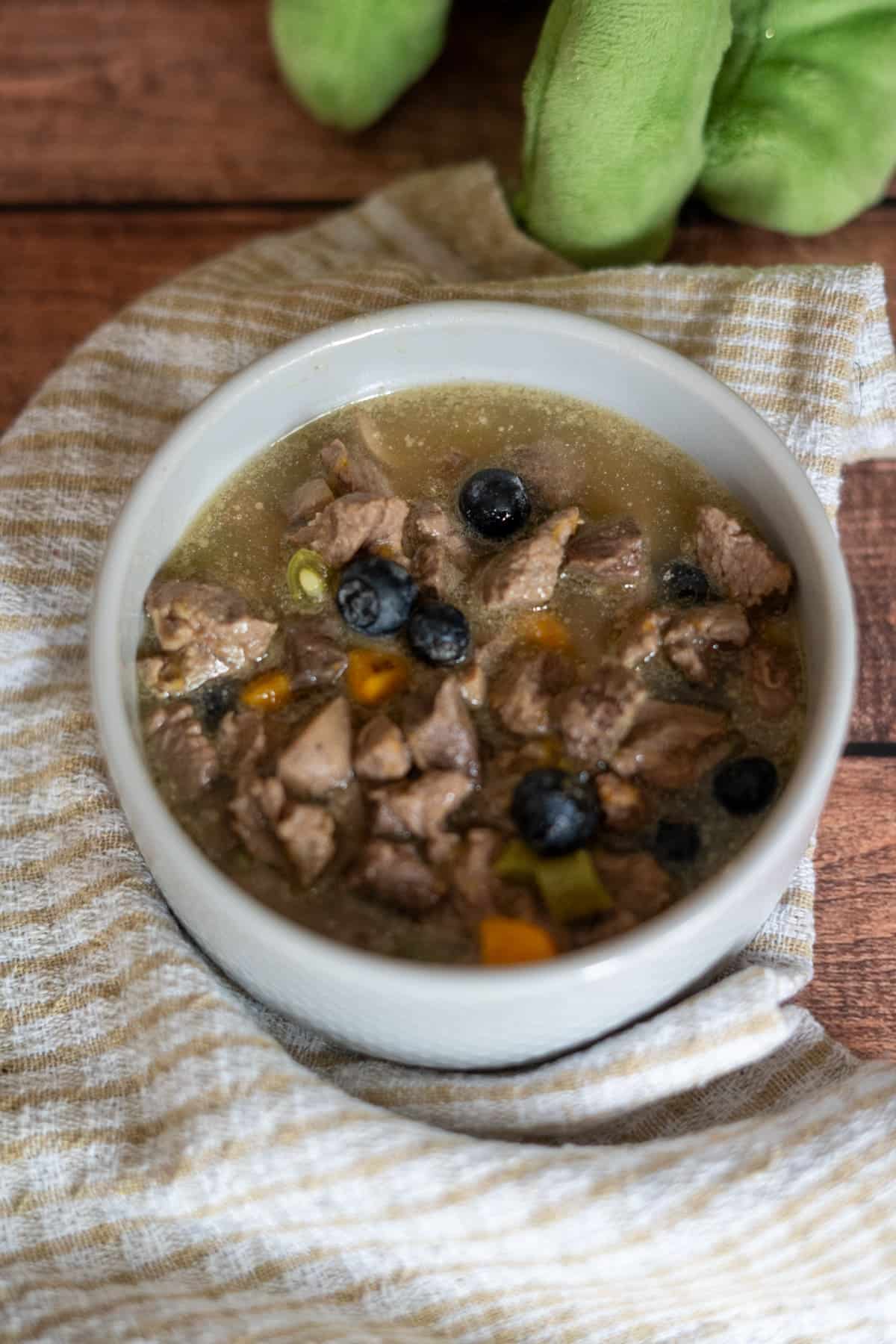 A white bowl filled with lamb stew for dogs, featuring chunks of meat, blueberries, and diced vegetables, placed on a striped cloth with a green plush object in the background.