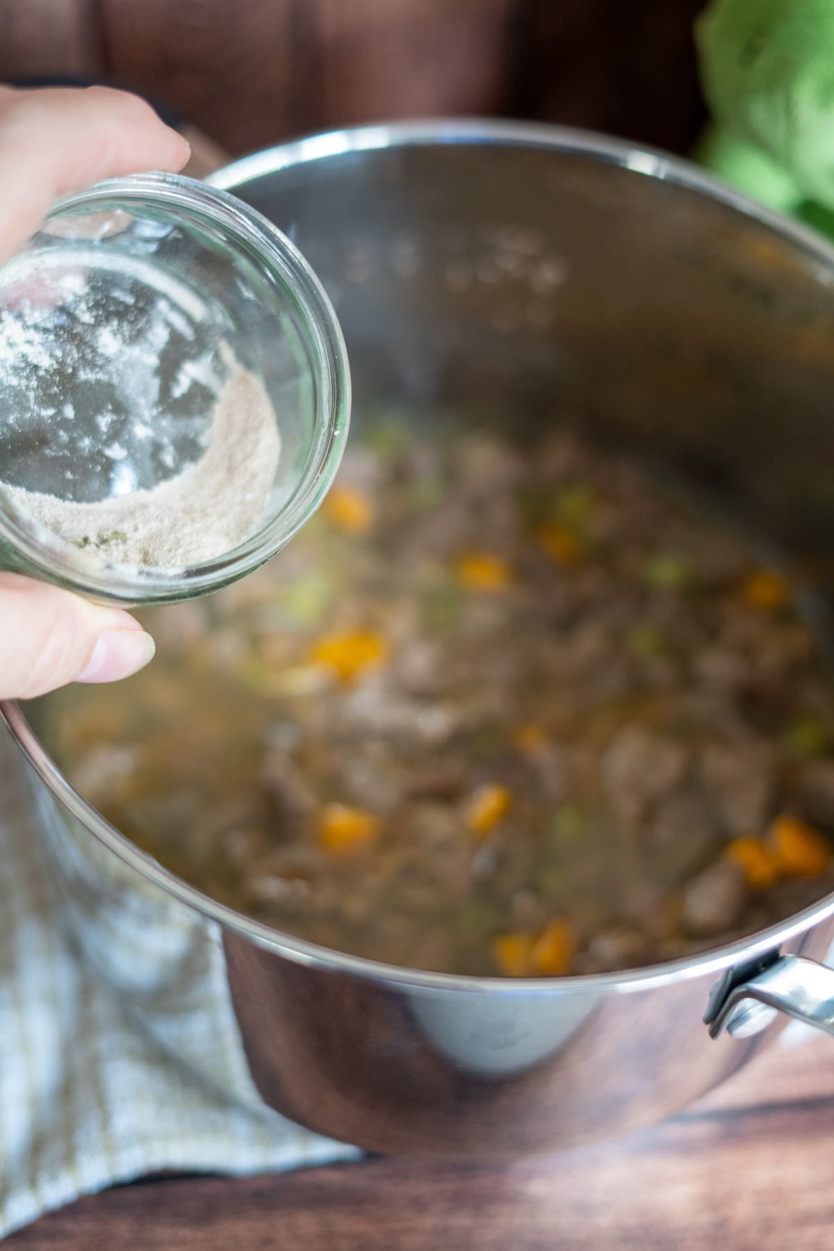 A hand sprinkles flour from a small glass bowl into a pot of lamb stew for dogs, filled with tender meat and vegetables on a hob.