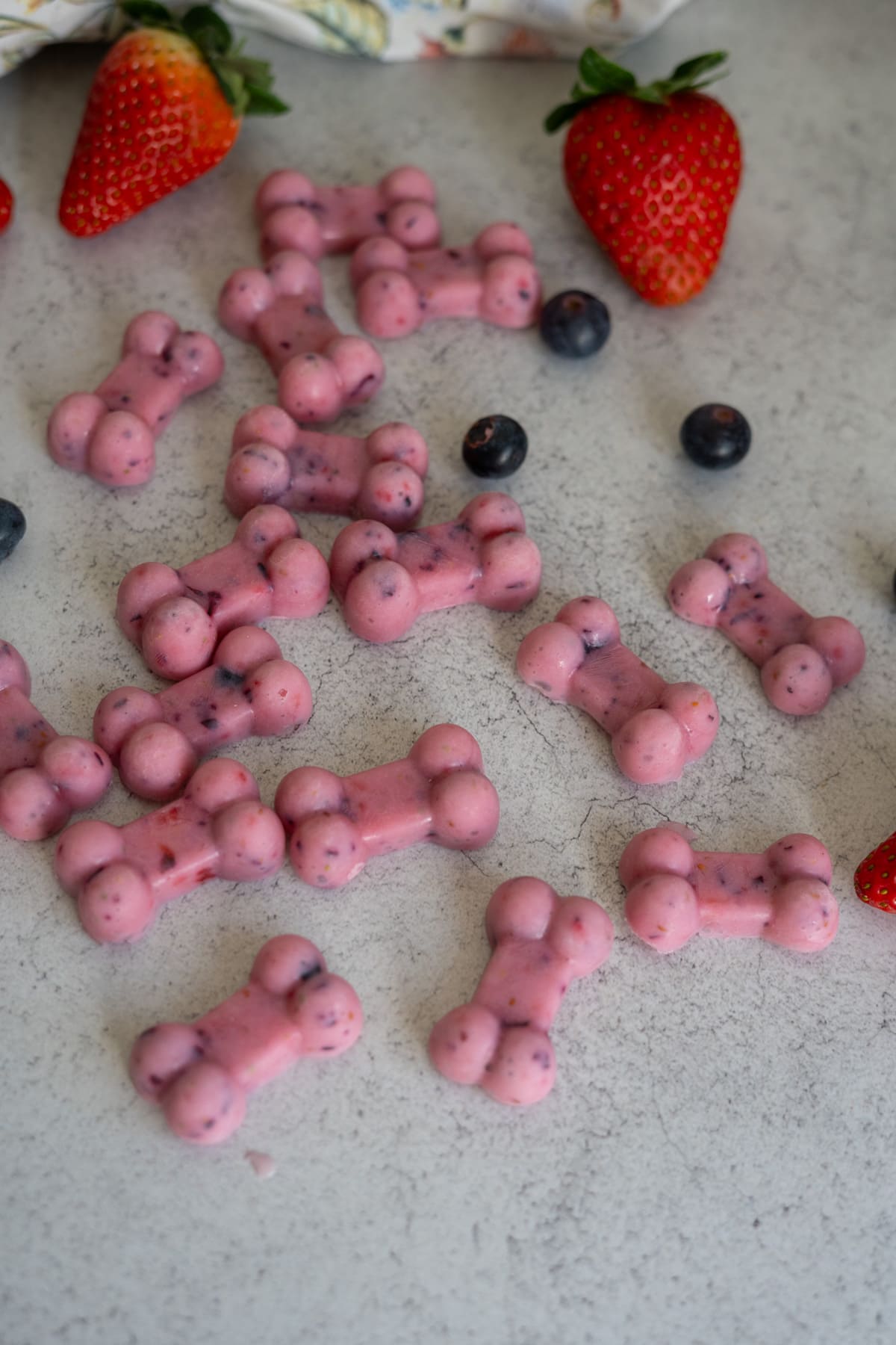 Several pink, bone-shaped kefir berry frozen dog treats with visible berry pieces are scattered on a light grey surface, surrounded by fresh strawberries and blueberries.
