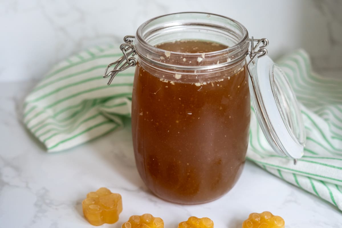 A glass jar filled with brown beef bone broth for dogs sits on a white surface near a green-striped cloth, with four small, flower-shaped yellow jellies in front.