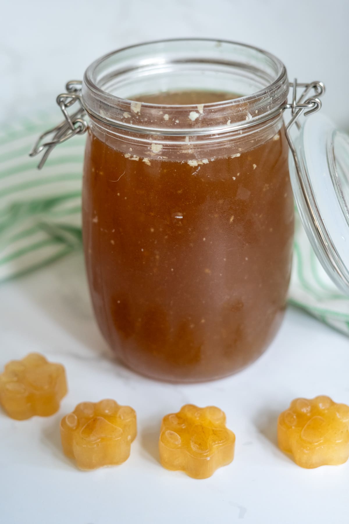 A glass jar filled with beef bone broth for dogs sits behind four small, paw-shaped gummy sweets on a white surface with a green-striped cloth in the background.