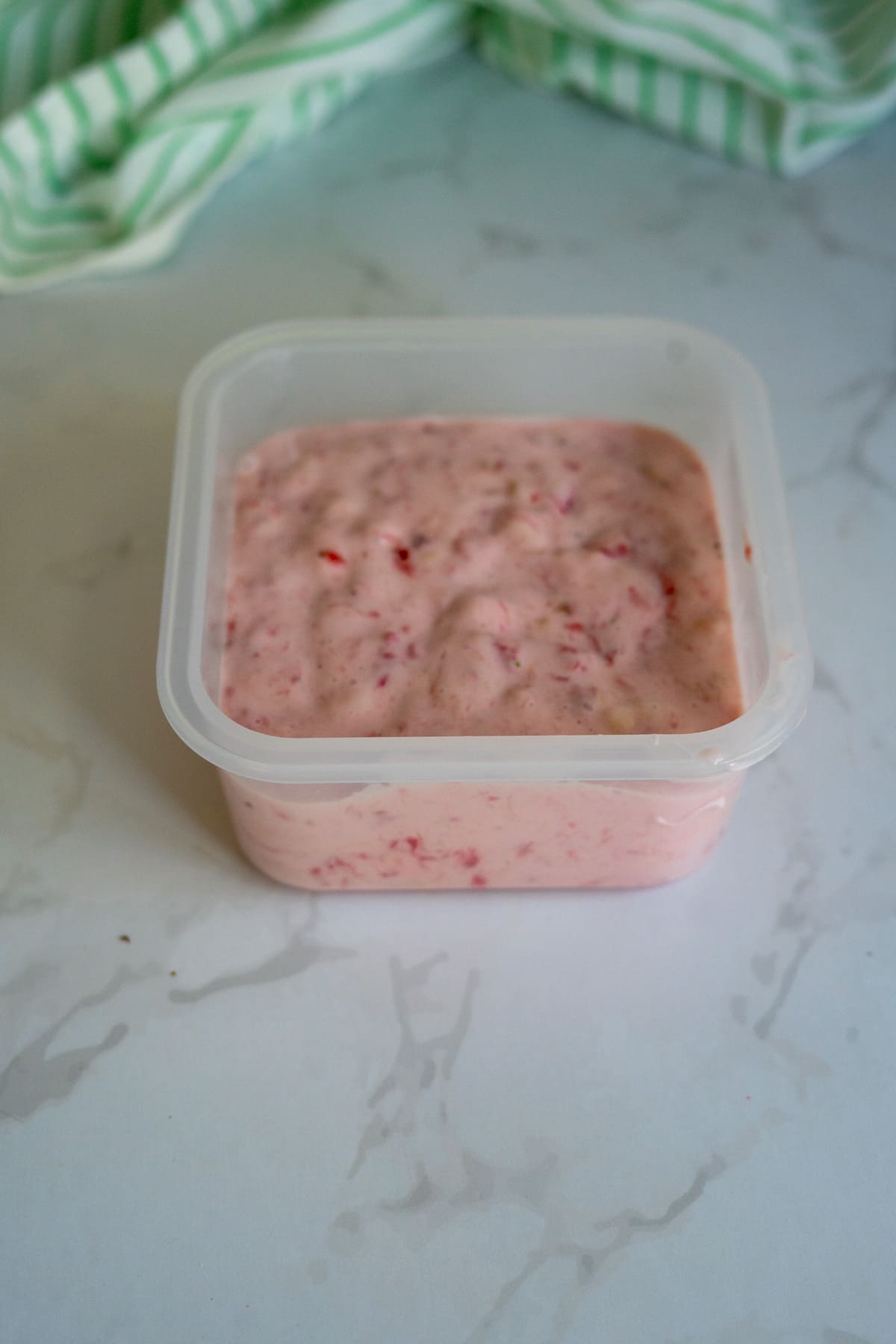 A plastic container filled with a pink, textured mixture sits on a light marble surface with a green and white striped cloth in the background.