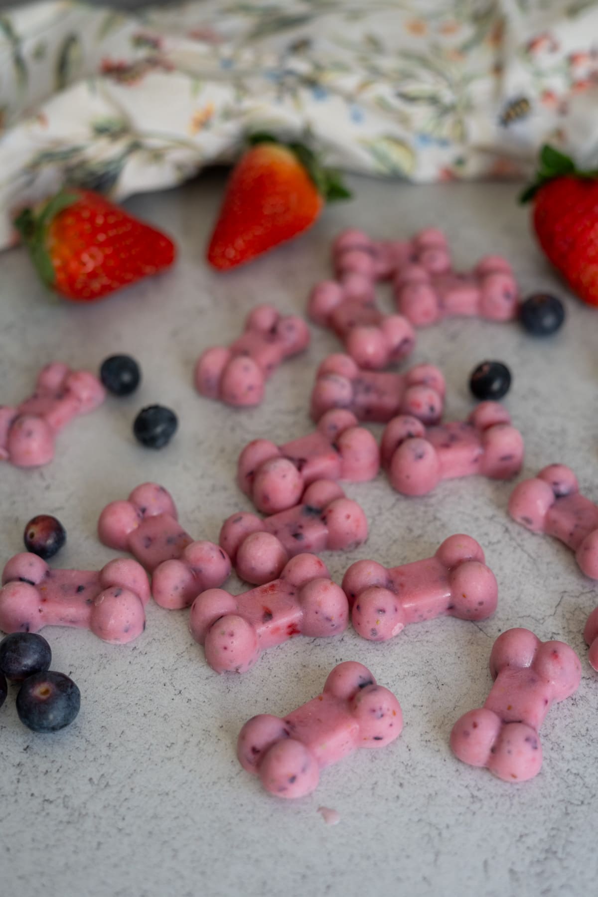 Pink, bone-shaped kefir berry frozen dog treats with visible berry pieces are scattered on a grey surface, surrounded by strawberries, blueberries, and a floral cloth.