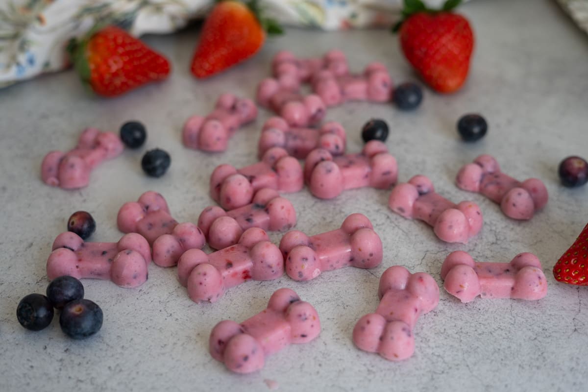 Pink bone-shaped treats made with berries are scattered on a grey surface, surrounded by whole strawberries and blueberries.
