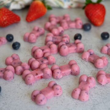 Pink bone-shaped frozen treats are scattered on a light surface, with strawberries and blueberries placed nearby.