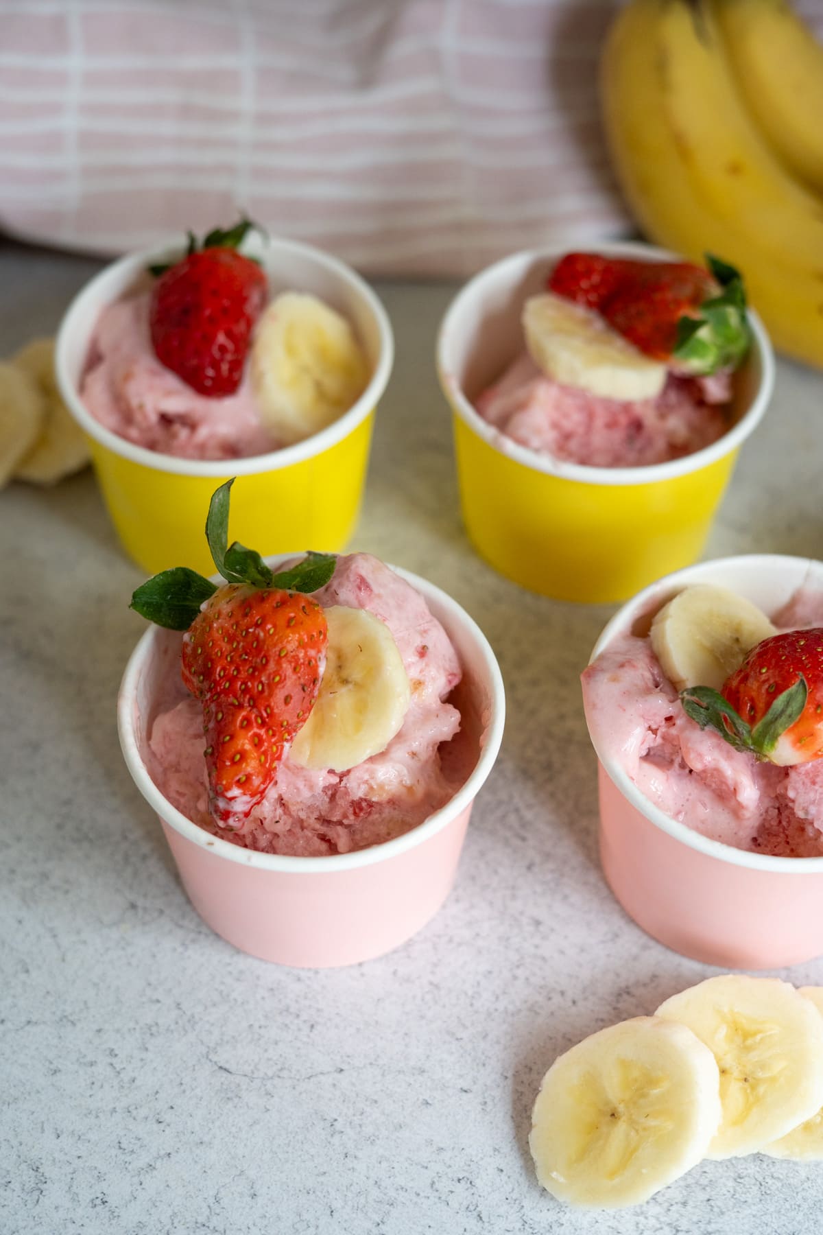 Four cups of pink strawberry-banana ice cream topped with fresh strawberry halves and banana slices, placed on a light surface with extra banana and strawberries nearby.