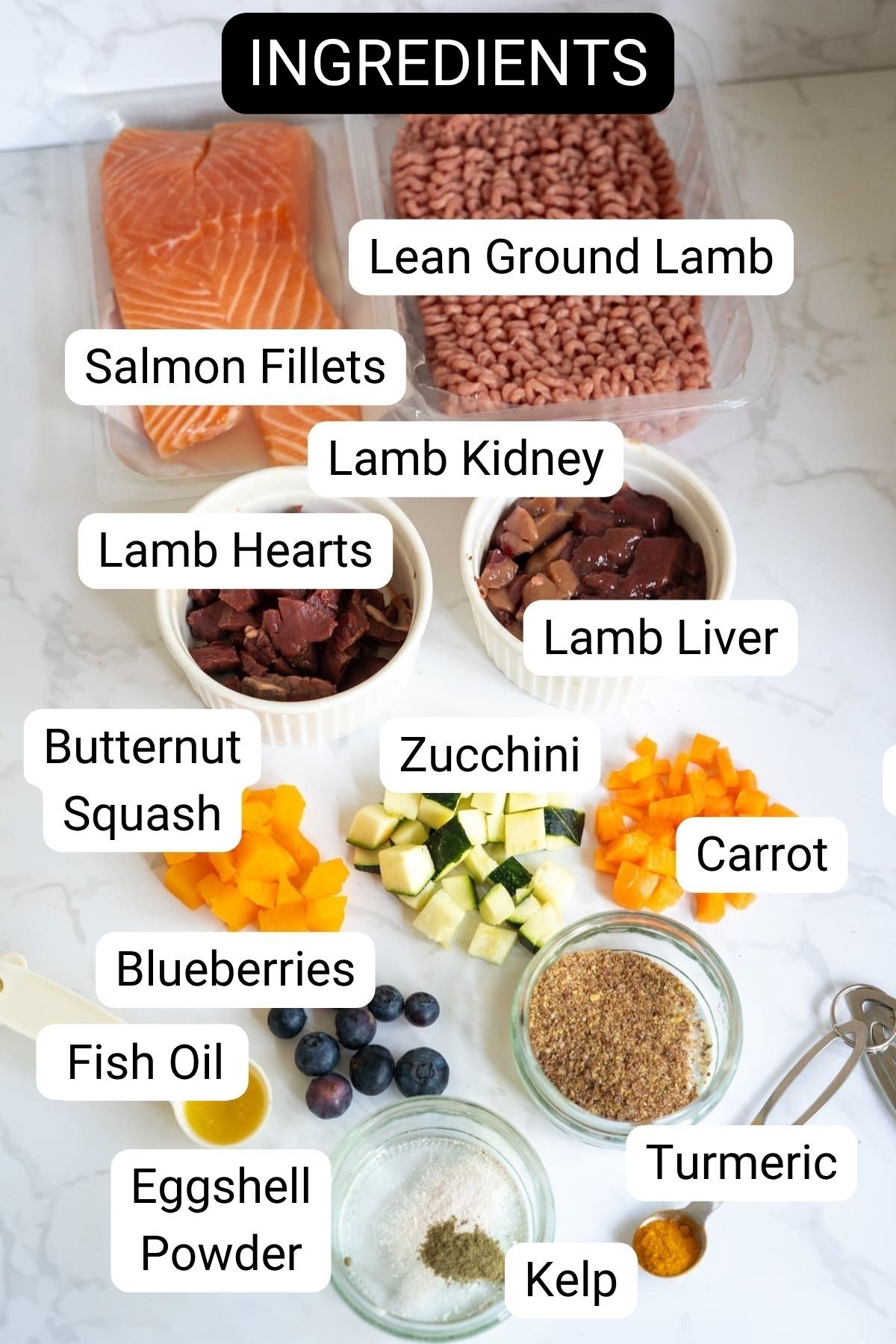 Various labelled ingredients on a counter, including salmon fillets, minced lamb, lamb kidney, lamb hearts, lamb liver, butternut squash, courgette, carrot, blueberries, fish oil, eggshell powder, turmeric and kelp.
