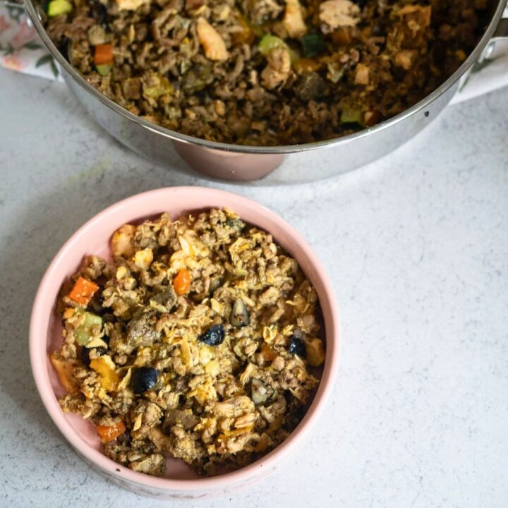 A pink bowl filled with a mixed dish containing vegetables, grains, and black beans sits next to a saucepan with more of the same mixture on a light worktop.