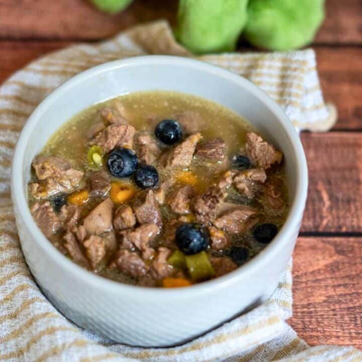 A white bowl filled with lamb stew for dogs, featuring chunks of meat, carrots, green vegetables, and blueberries, sits on a striped cloth atop a wooden surface.