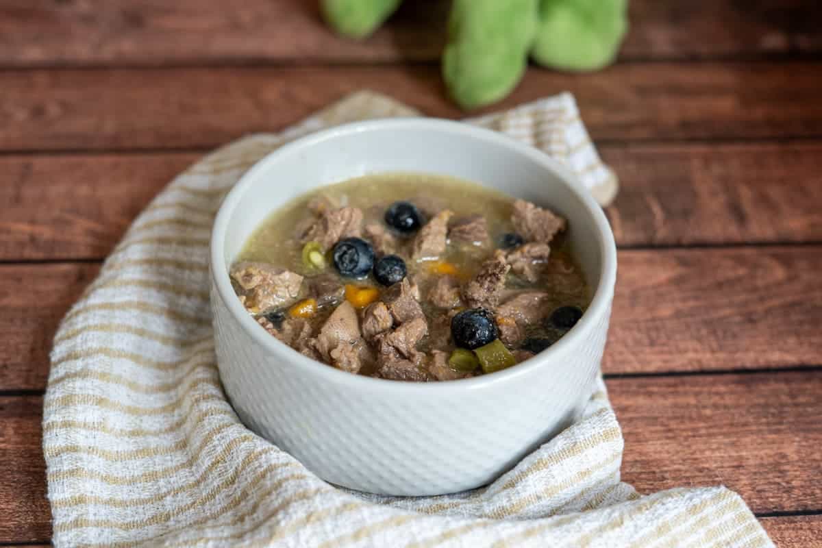 A white bowl filled with lamb stew for dogs, mixed with vegetables and topped with blueberries, sits on a striped cloth on a wooden surface.
