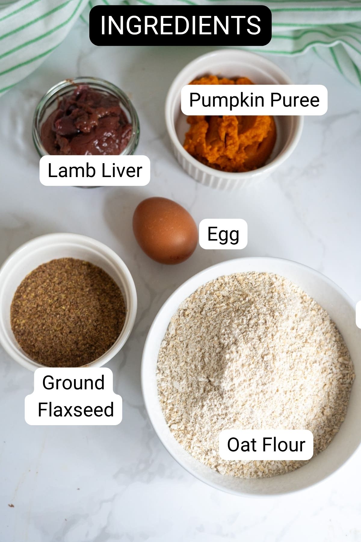 Bowl of oat flour, ground flaxseed, pumpkin purée, lamb liver, and a whole egg arranged on a white surface; each ingredient is labelled.