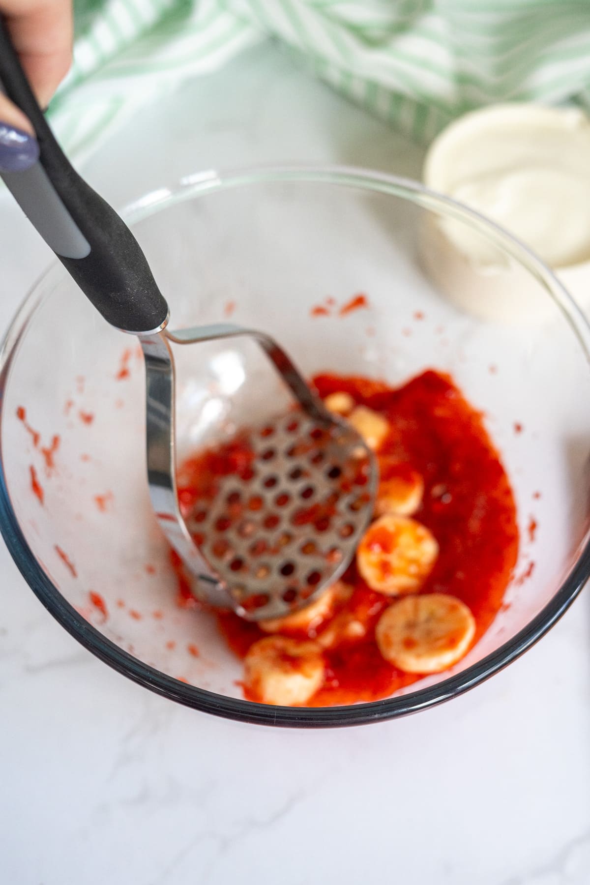 A potato masher mashing sliced bananas and strawberries in a glass bowl on a white surface.