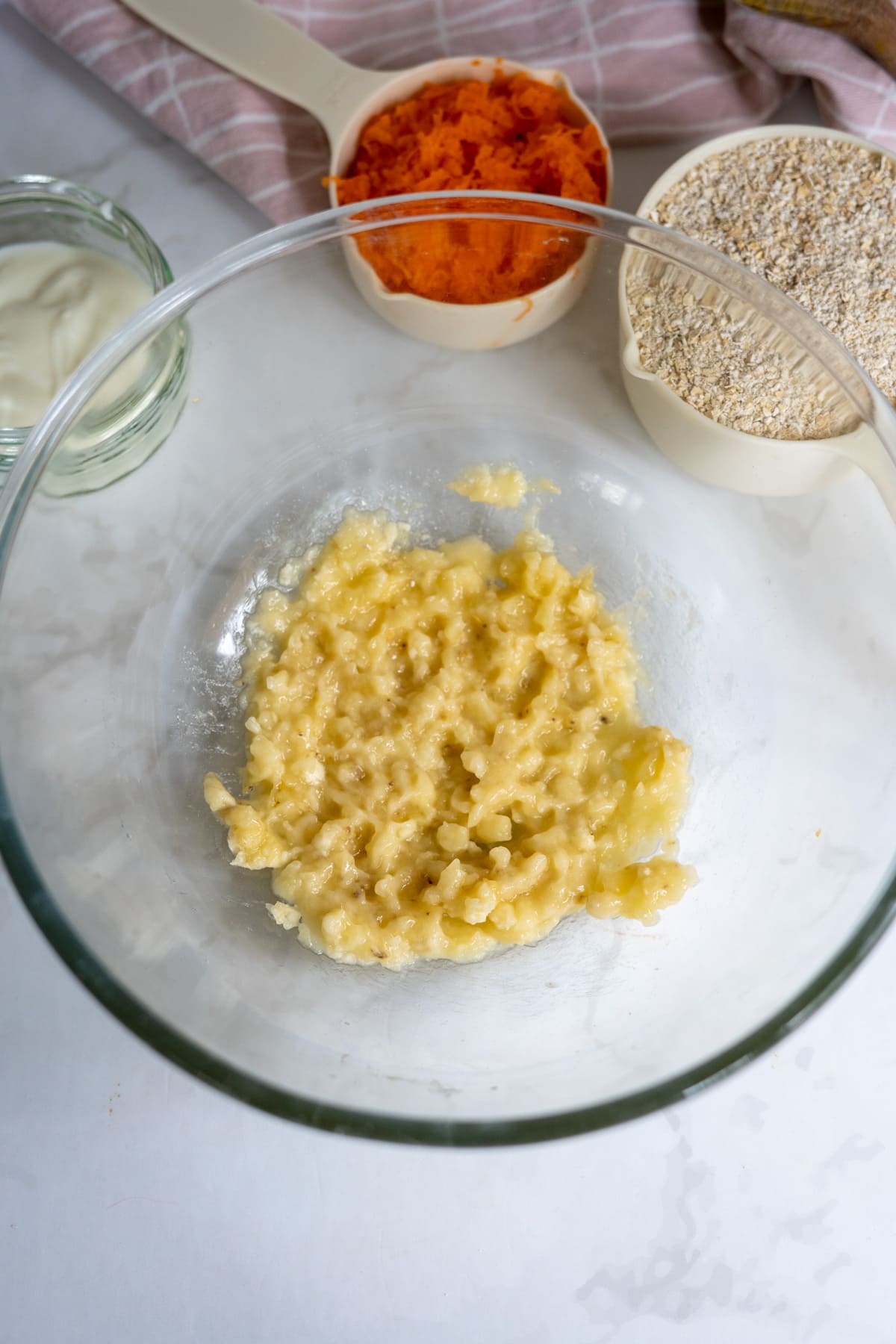 A glass bowl with mashed bananas sits on a white surface, surrounded by small bowls of oats, yoghurt, and grated carrots—perfect ingredients for homemade banana carrot dog treats.