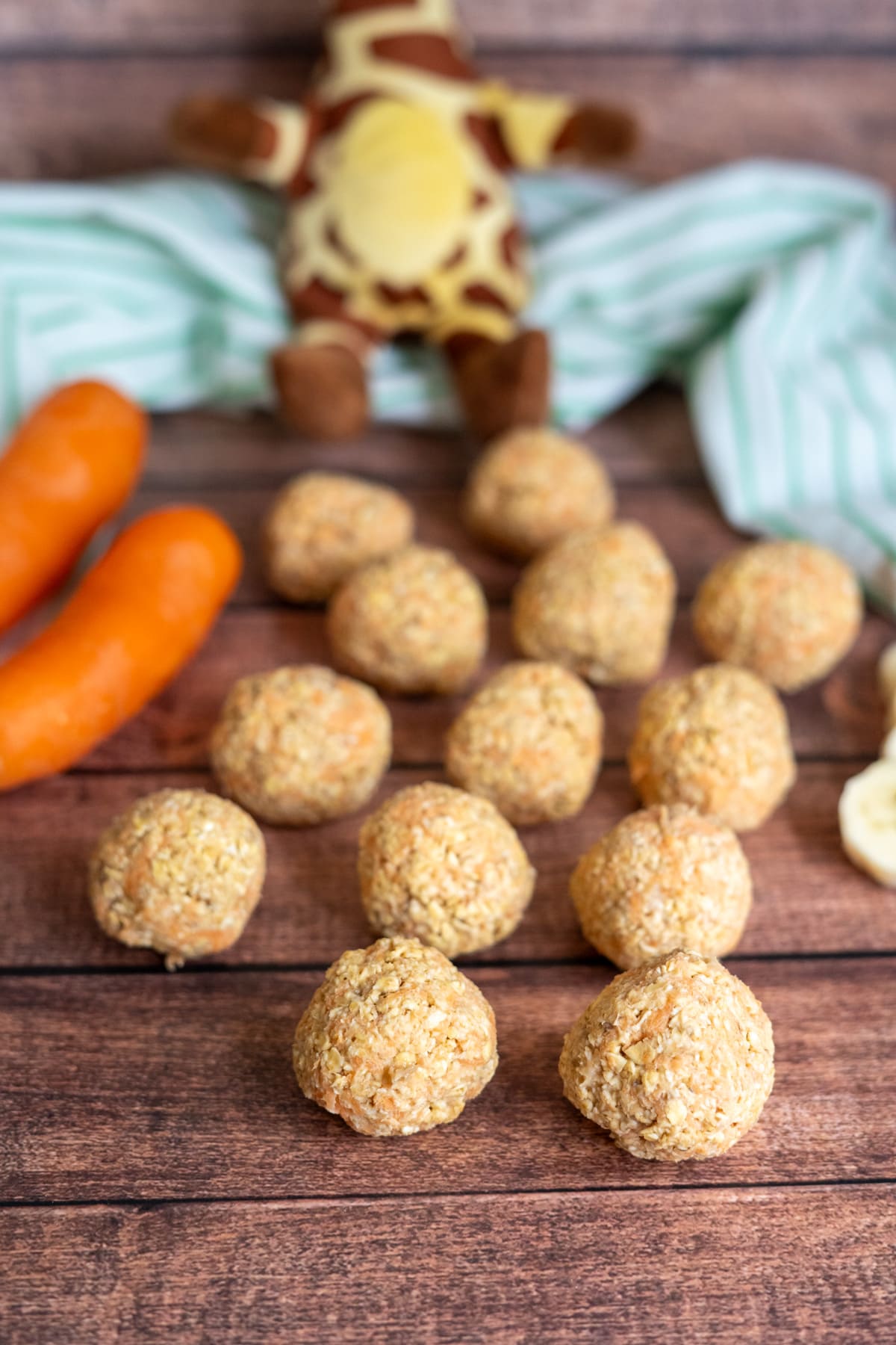 Several oat balls, crafted as banana carrot dog treats, are arranged on a wooden surface with two carrots, banana slices, a striped cloth, and a plush giraffe in the background.