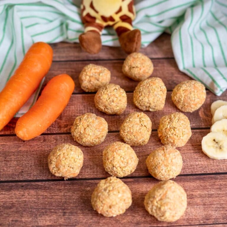 Round oat balls are arranged on a wooden surface next to two whole carrots, banana slices, and a striped cloth with a soft toy giraffe partially visible in the background.