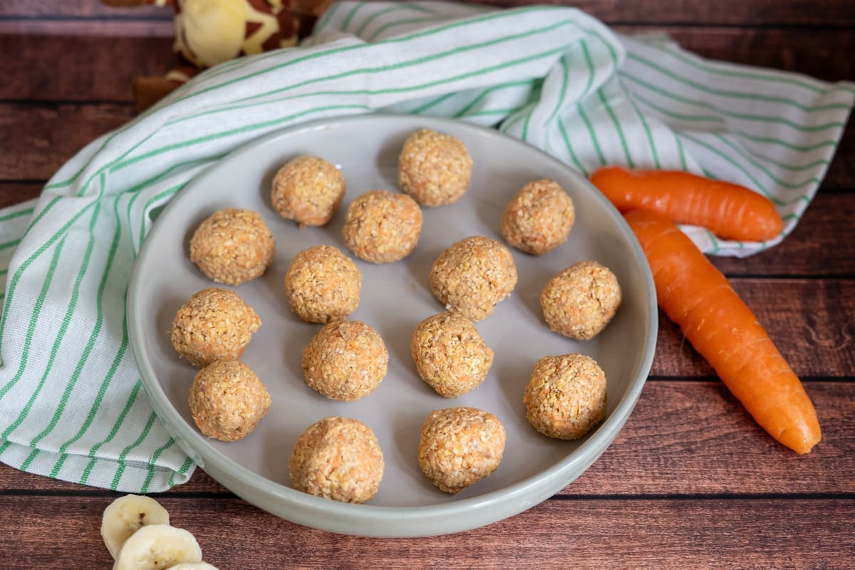 A plate of round, homemade snack balls sits on a striped cloth, with two whole carrots and banana slices nearby on a wooden surface.