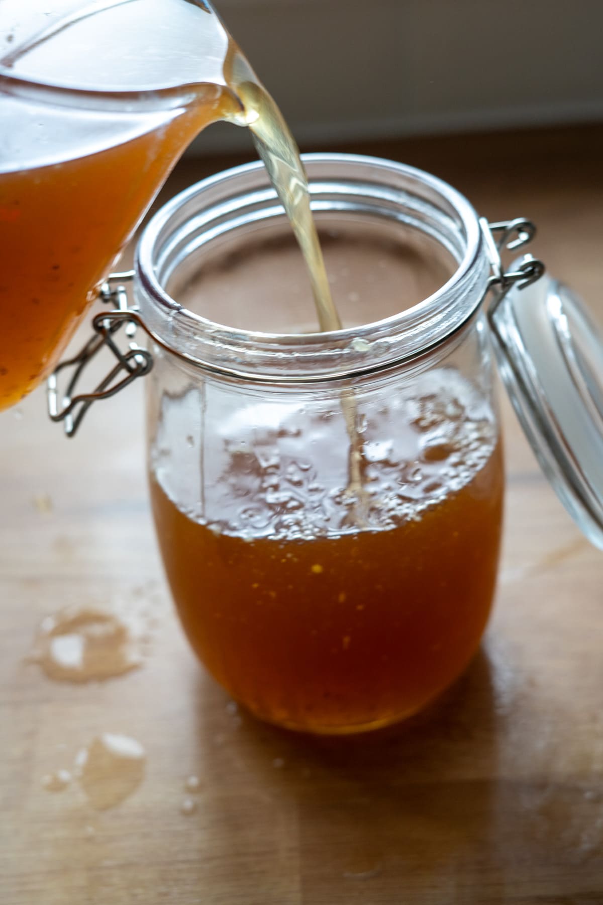 A jar is being filled with golden brown beef bone broth for dogs from a jug, on a wooden surface with some spilt drops nearby.