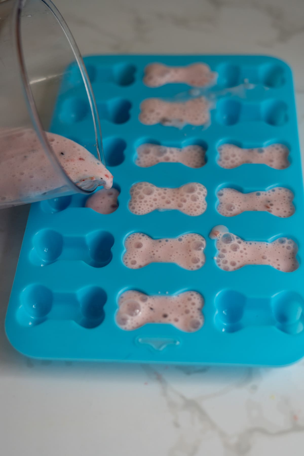 A person pours a pink, foamy mixture into a blue silicone mould with bone-shaped cavities on a marble surface.