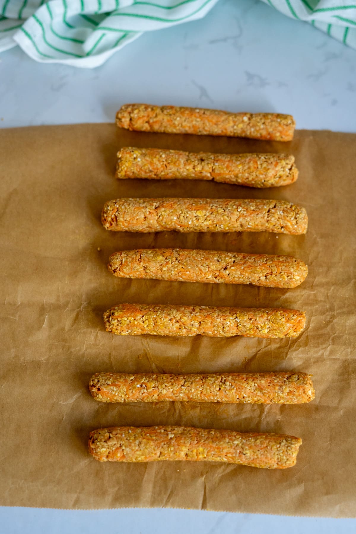 Eight homemade duck dog treats in stick shapes are arranged in a row on brown baking parchment, with a green and white striped cloth in the background.