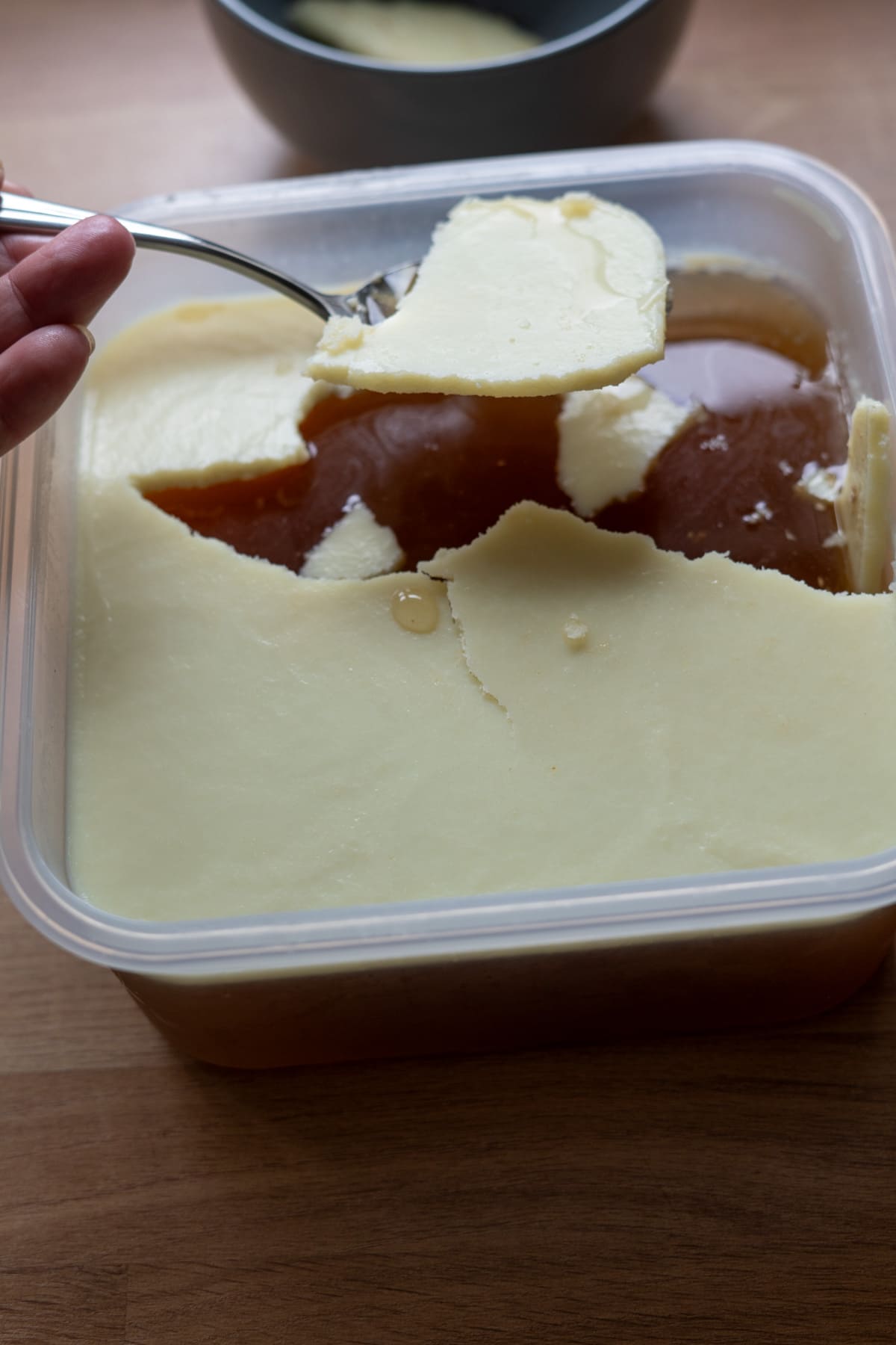 A hand holds a spoon lifting a layer of solidified fat from the surface of beef bone broth for dogs in a plastic container on a wooden surface.