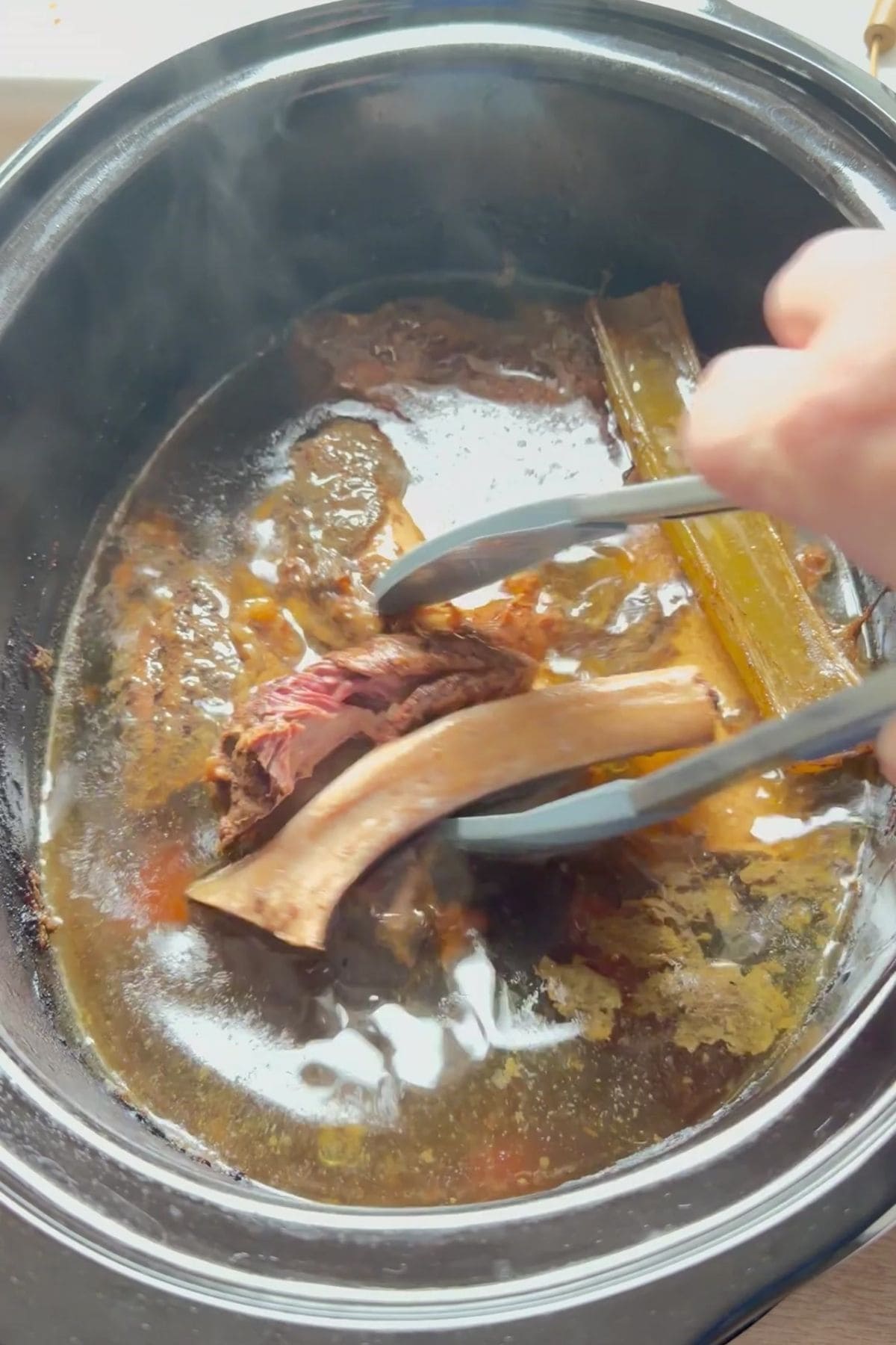 A hand uses tongs to lift a cooked beef short rib from a slow cooker filled with vegetables, bones, and nourishing beef bone broth for dogs.