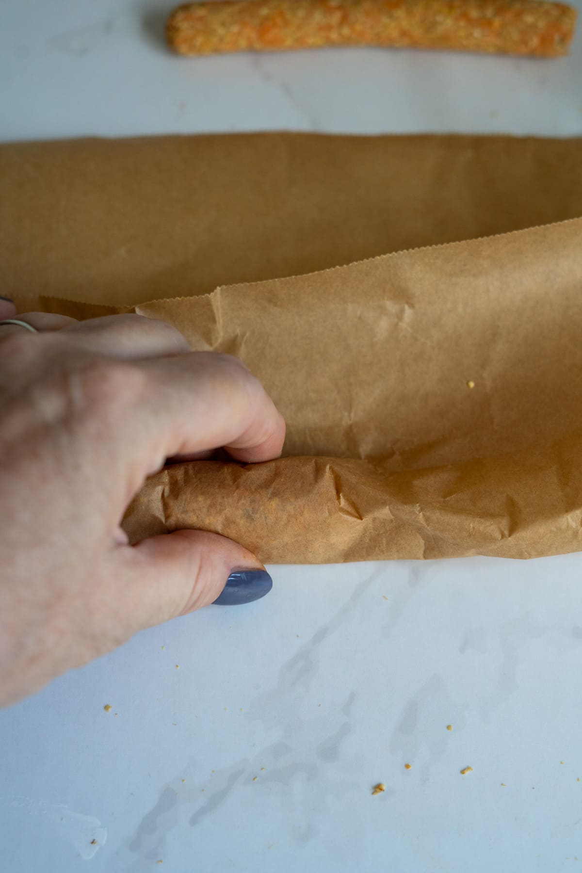 A hand is folding a sheet of brown greaseproof paper on a white surface, with homemade duck dog treats rolled in pastry in the background.