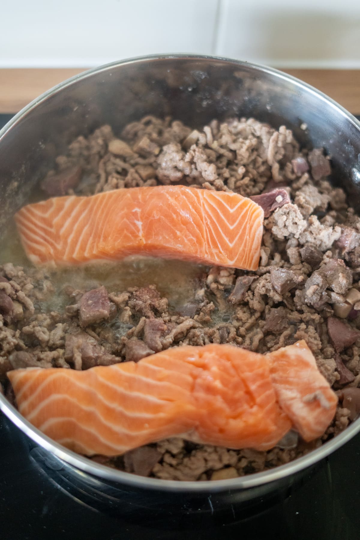 Two raw salmon fillets placed on top of cooked minced meat and offal pieces in a stainless steel saucepan on a hob.