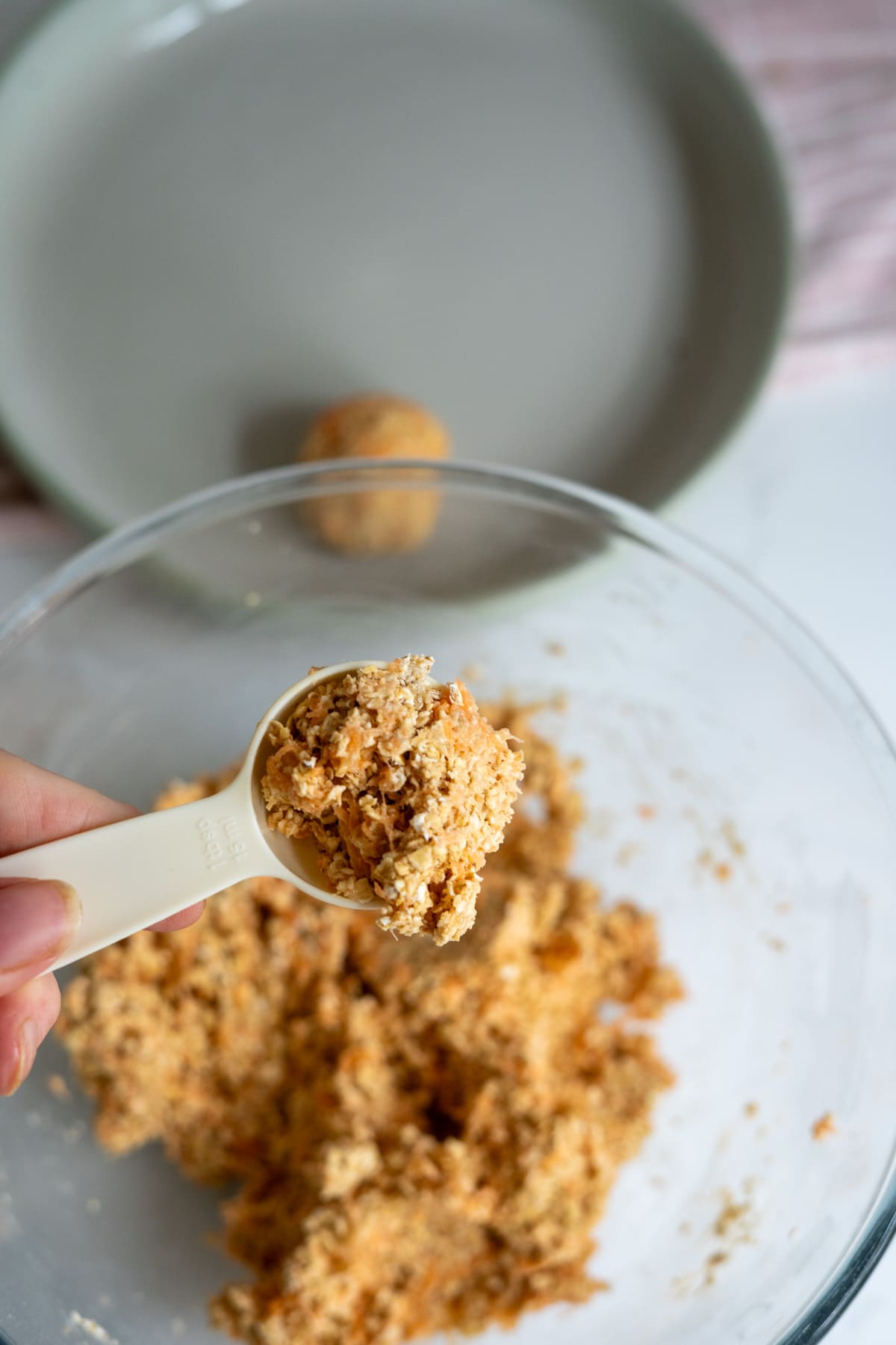 A hand holds a tablespoon of crumbly banana carrot dog treats mixture over a glass bowl, with a formed ball and a plate in the background.