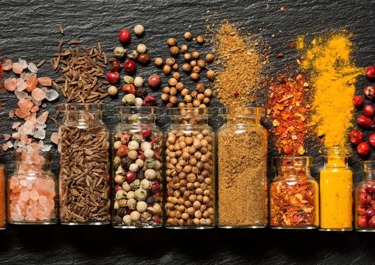 Glass jars filled with various spices and seasonings, including peppercorns, seeds, powders, and pink salt, arranged in a row on a dark slate surface.