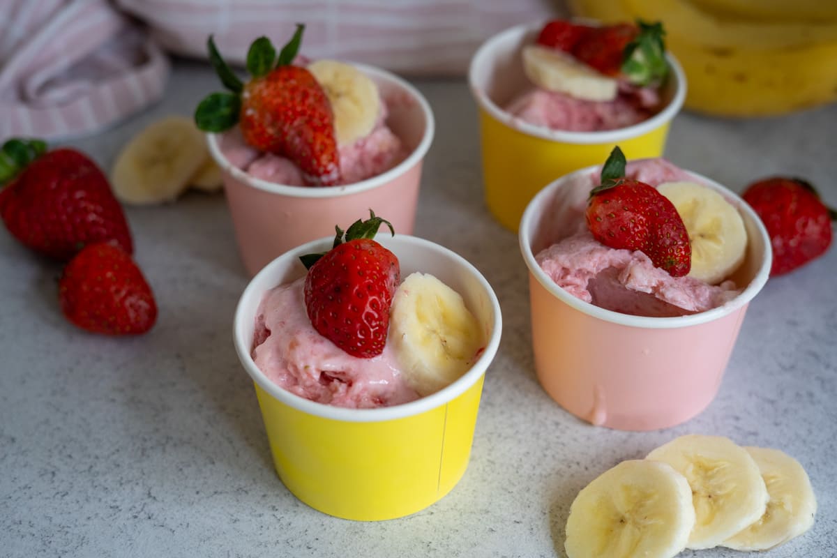 Four bowls of pink ice cream topped with strawberries and banana slices, with extra fruit placed on a light-coloured surface.