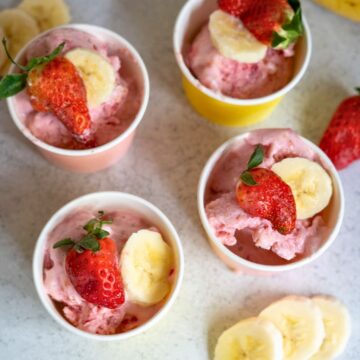 Four small pots of pink ice cream are topped with fresh strawberries and banana slices, with extra strawberries and banana slices on the white surface nearby.