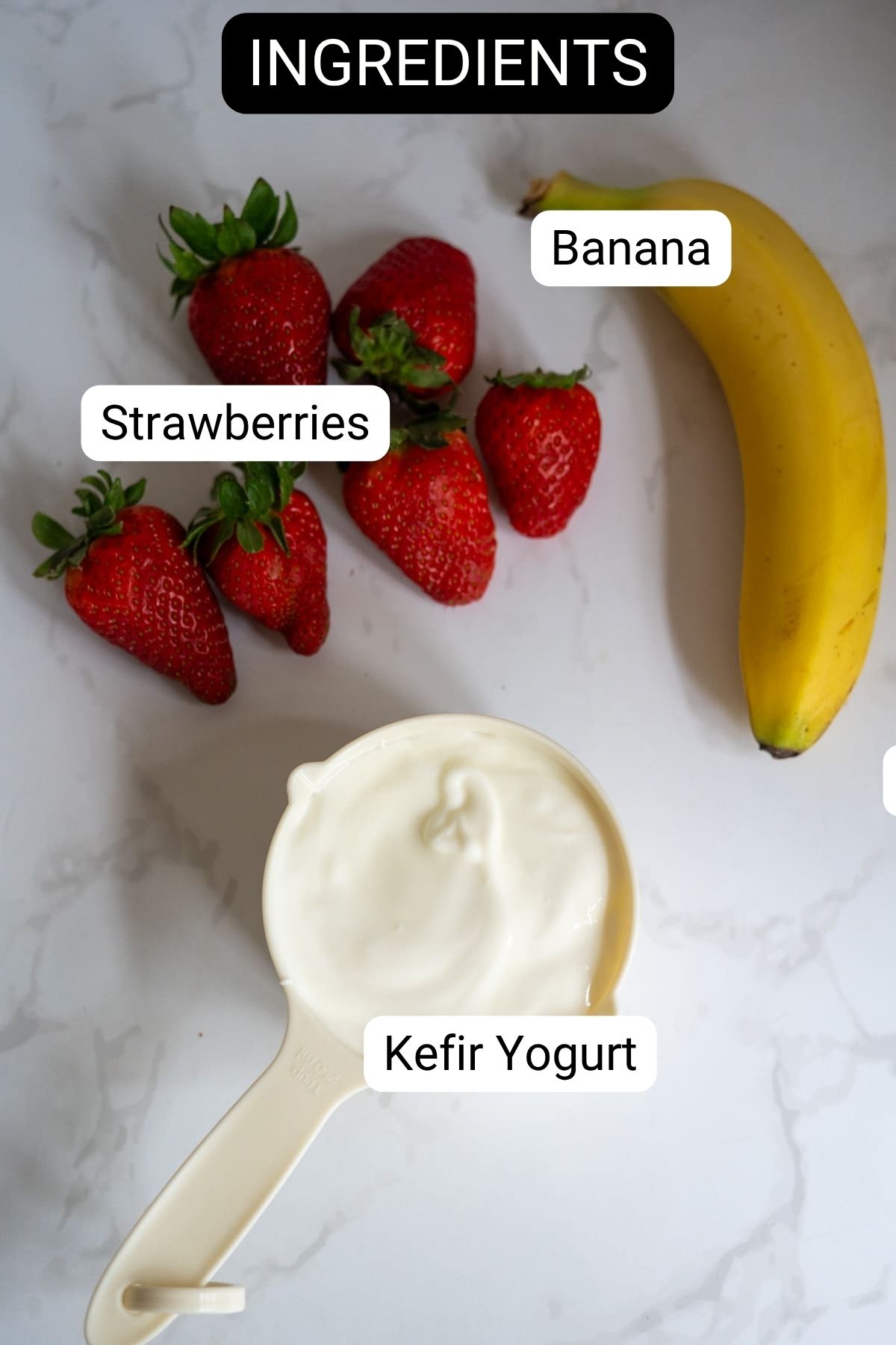 A banana, strawberries, and a measuring jug of kefir yoghurt are arranged on a white surface, each labelled with its name.