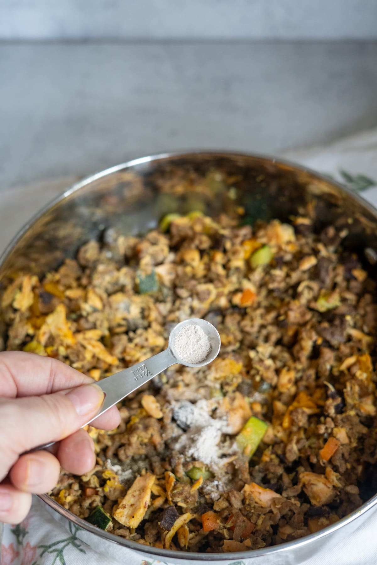A hand holds a measuring spoon of white powder over a stainless steel pan filled with a mixed vegetable and meat stir-fry.