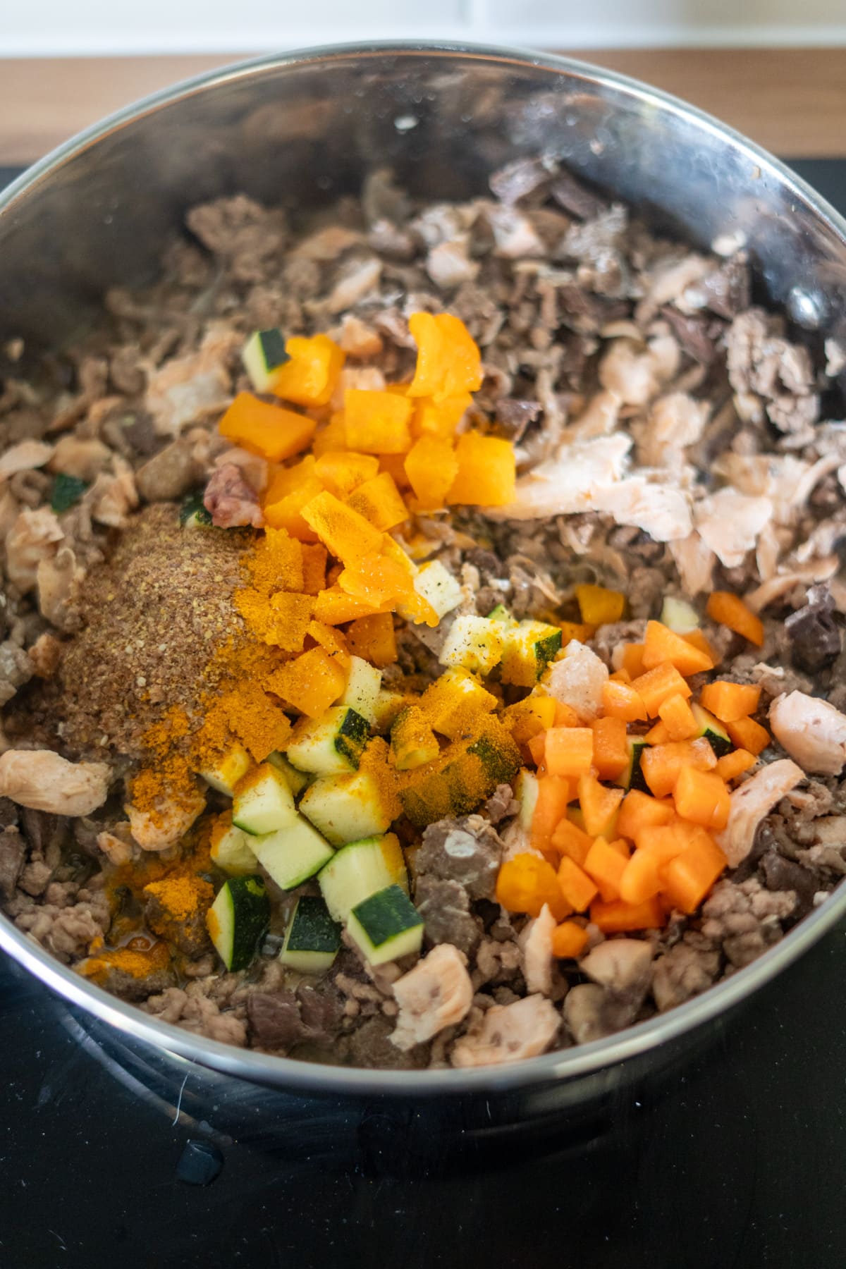 A stainless steel saucepan on a hob filled with chopped carrots, courgette, orange pepper, minced meat, and spices.