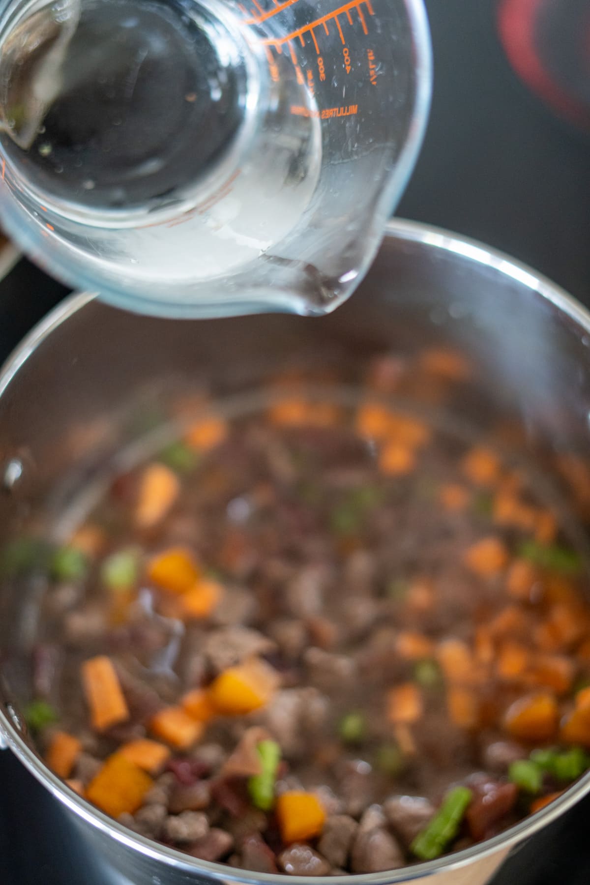 A glass measuring jug pours liquid into a saucepan containing diced meat, carrots, green beans, and other ingredients for a wholesome lamb stew for dogs.