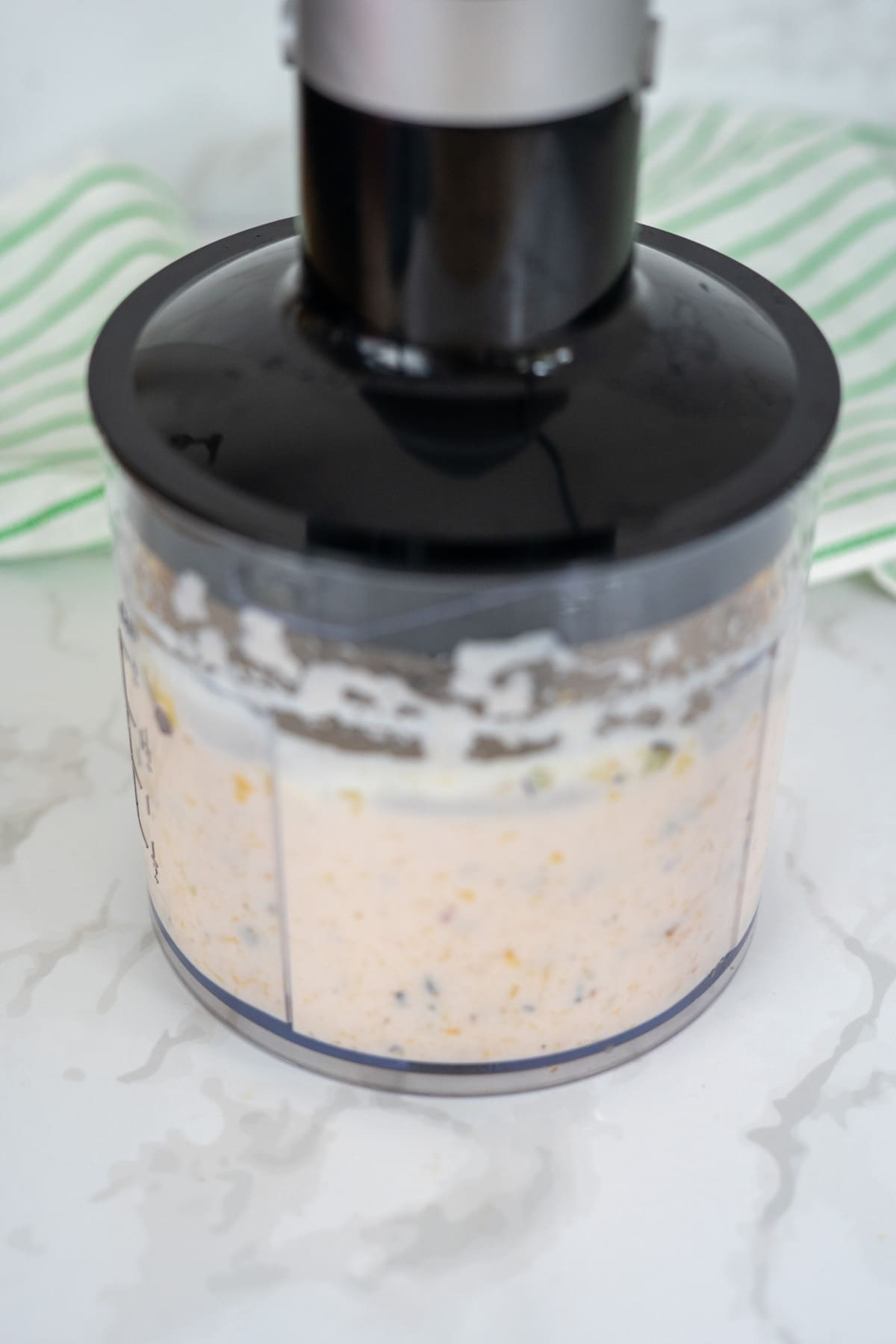 A food processor blending a creamy mixture with visible small pieces, placed on a white marble surface with a green-striped cloth in the background.