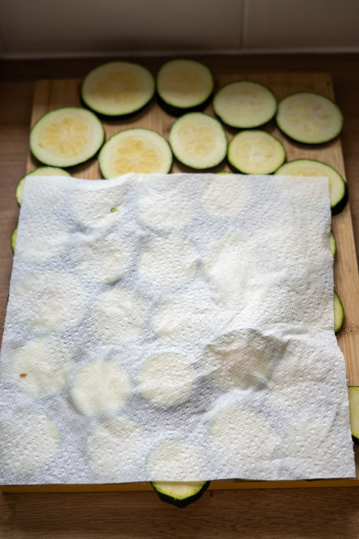 Sliced courgette rounds on a wooden chopping board, partially covered with a sheet of white kitchen roll.