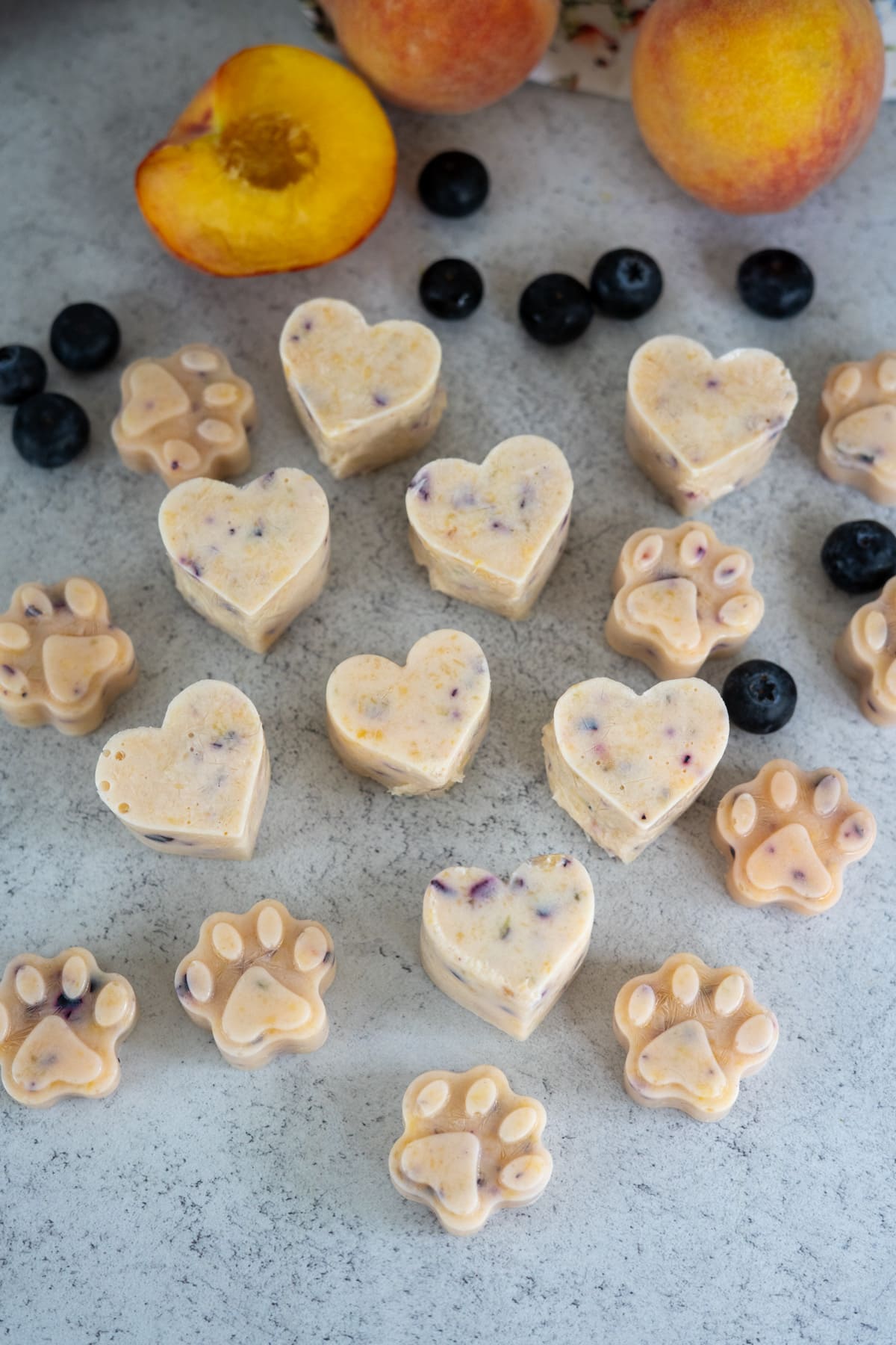 Heart- and paw-shaped frozen peach and blueberry dog treats with visible fruit pieces are arranged on a worktop, surrounded by fresh blueberries and whole peaches.