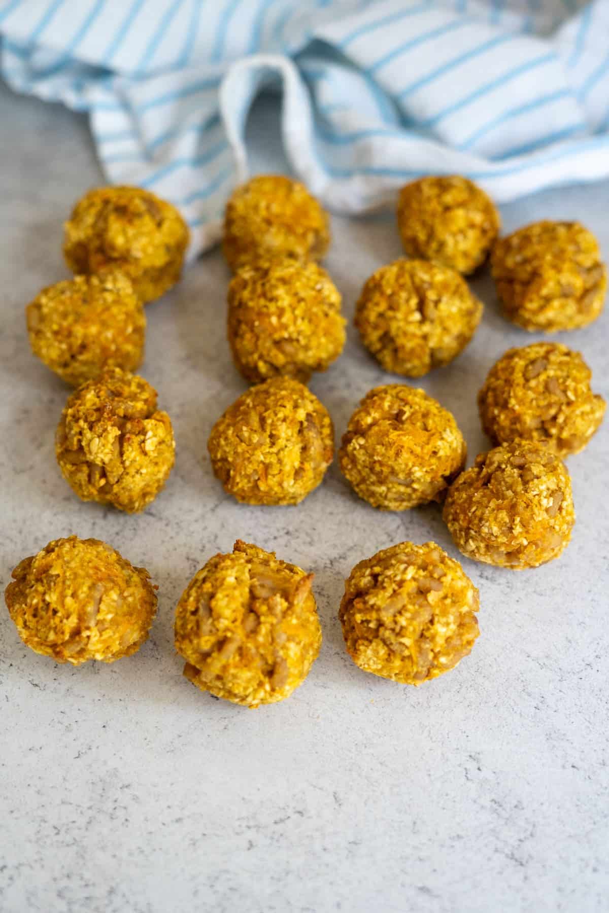 Fifteen round, orange-coloured oat balls are arranged on a light grey surface with a blue and white striped cloth in the background.