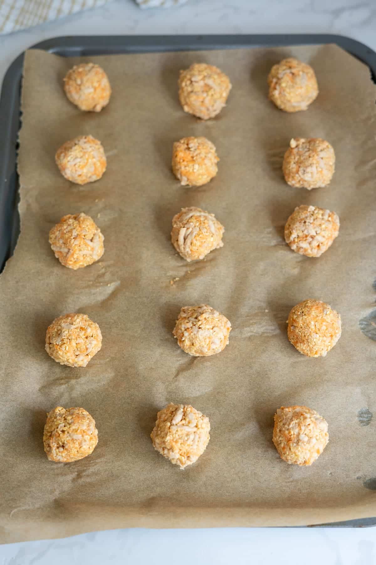 Baking tray lined with baking paper holds fifteen evenly spaced, uncooked, round dough balls.