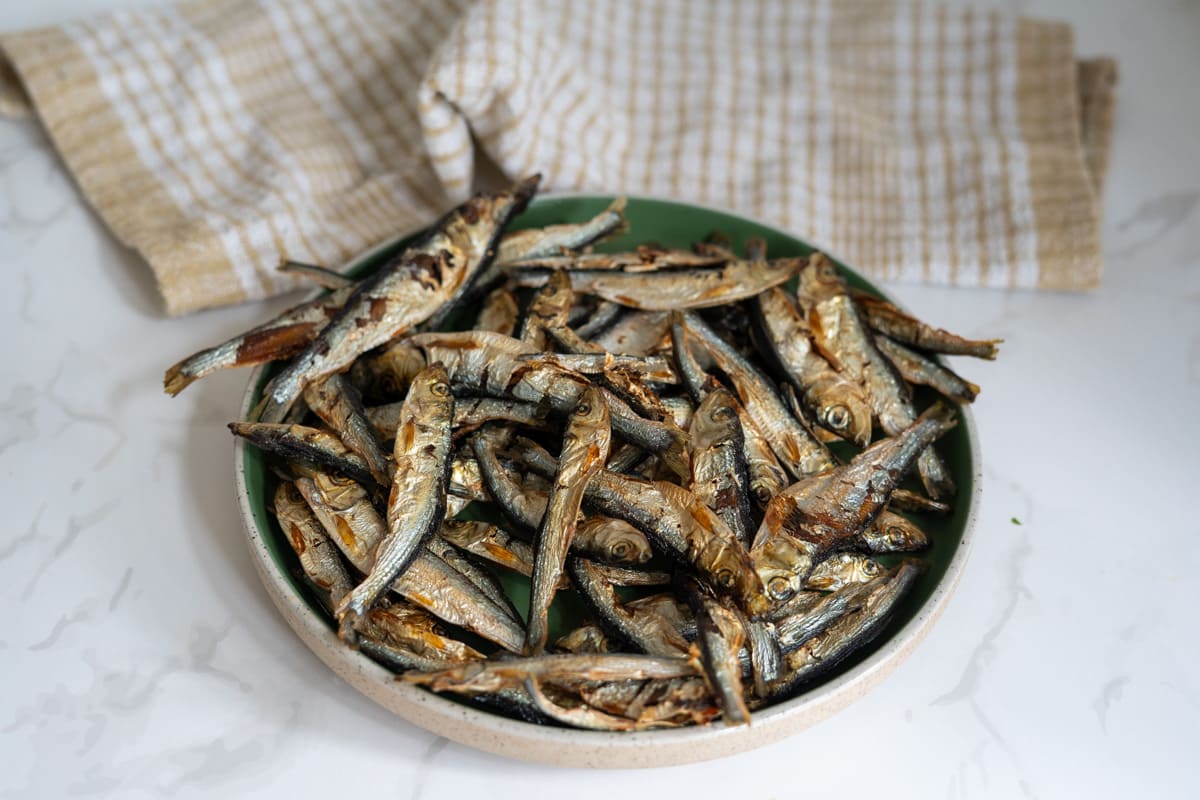 A green plate filled with dried fish sits on a white surface, with a beige and white checked tea towel in the background.