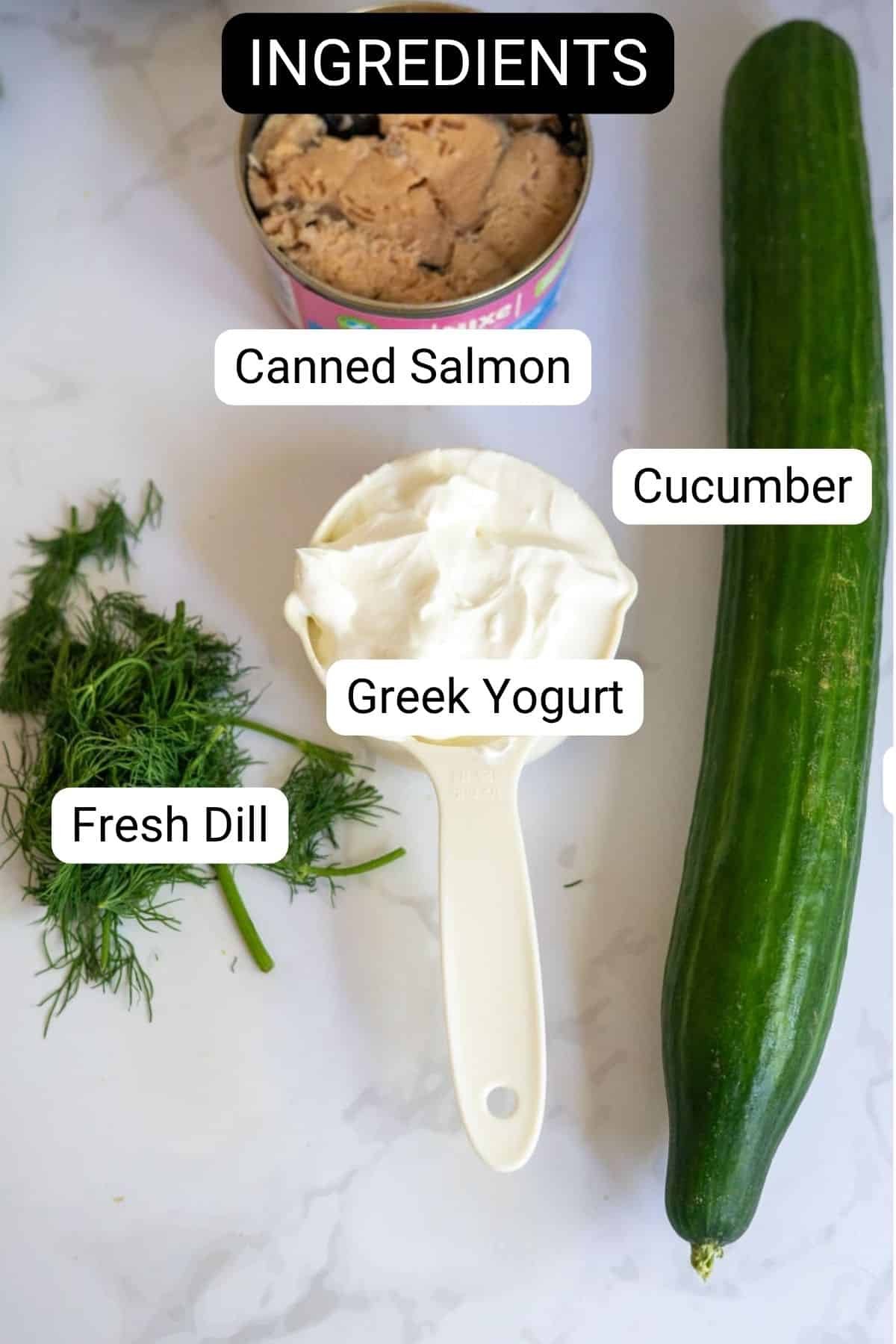 Tinned salmon, a cucumber, fresh dill, and a measuring jug of Greek yoghurt are arranged on a white surface, each ingredient labelled.