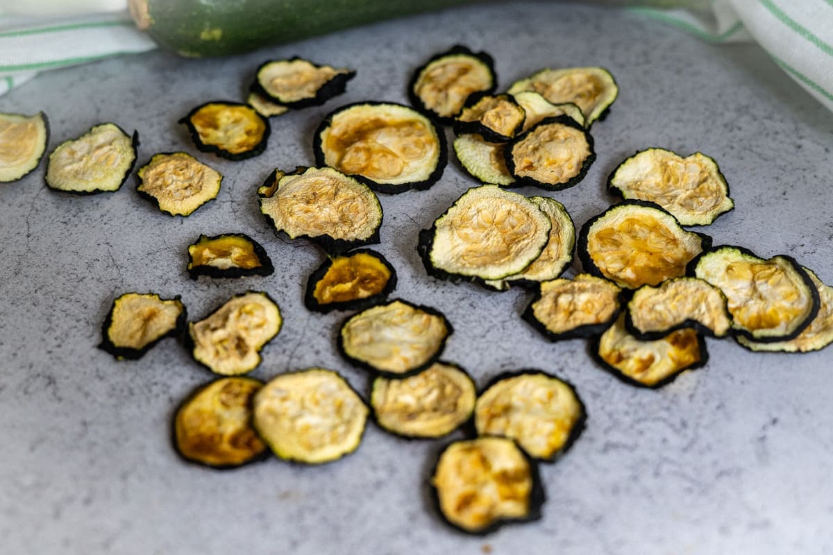 Slices of dried courgette are scattered on a grey worktop, with part of a fresh courgette visible in the background.