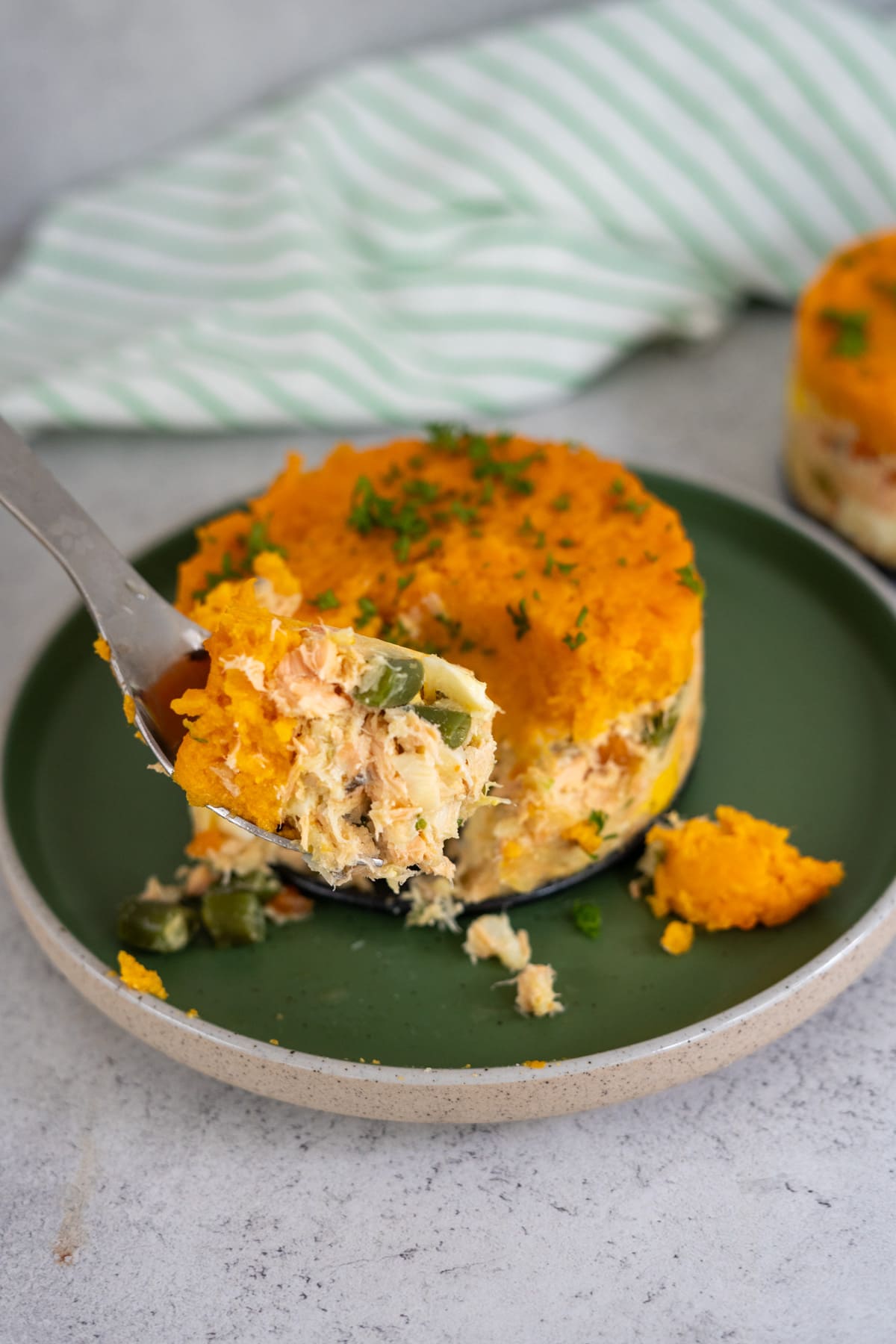A fork holds a bite of baked casserole topped with mashed sweet potatoes, served on a green plate, with a striped cloth in the background.