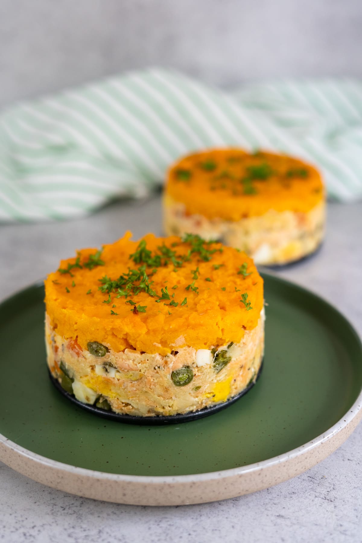Two individual servings of fish pie for dogs, layered with mashed sweet potato on top and a mix of vegetables and tender fish beneath, garnished with chopped herbs on a green plate.