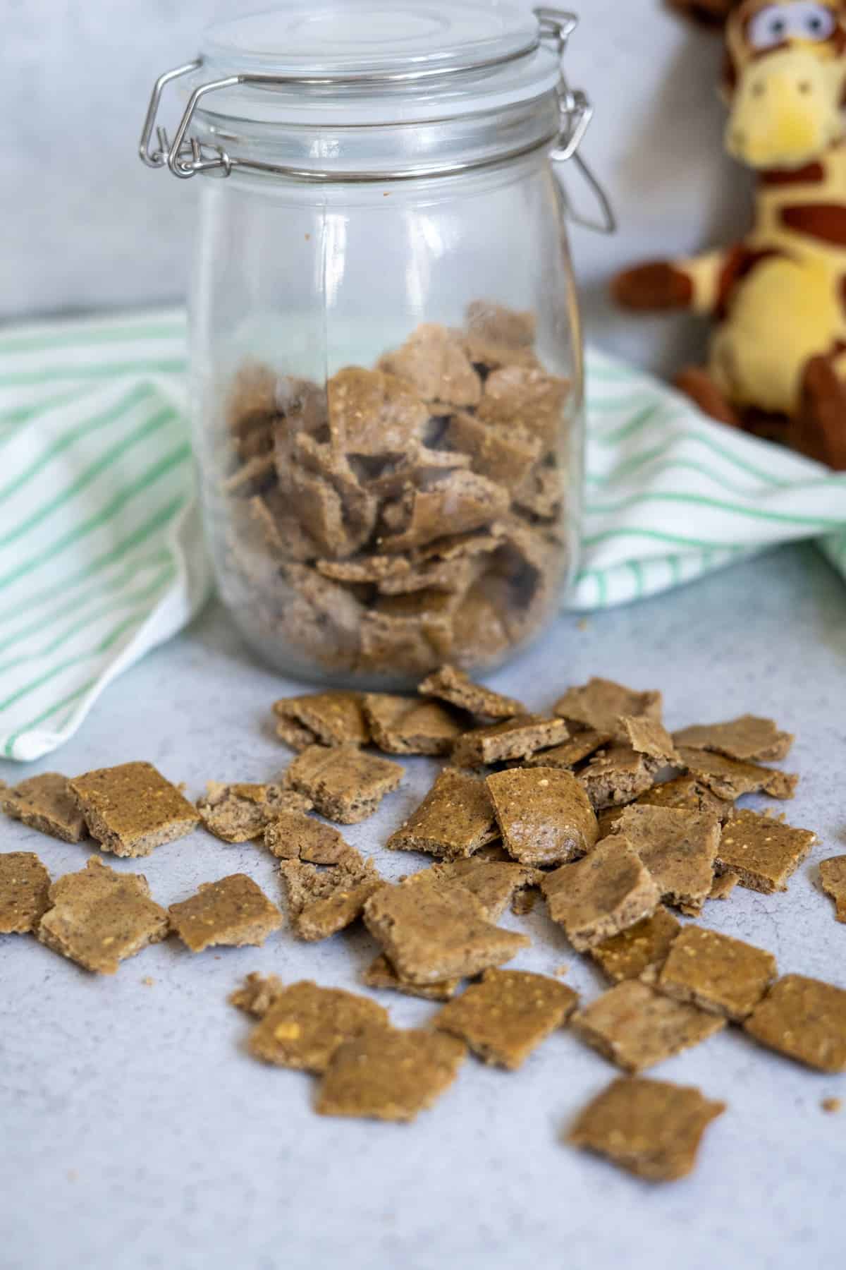 A glass jar filled with square-shaped homemade duck dog treats sits on a counter, with more treats scattered in front and a stuffed giraffe toy in the background.